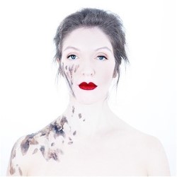 A promotional photo for the performance of a white woman, powdered to look even whiter, with dark hair pulled back and red lipstick. She has no expression and stares straight ahead. On her face and down her right side are splashes of dark mud or paint.