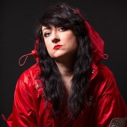 Promotional photo of Bee Babylon, a white woman with long black hair looking moody and serious in a red raincoat.