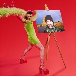 Promotional photo of Simon David. He is posing in a full-body shot in pink heels, a green minidress and a green boa with his feet apart and arms thrown wide. He is beside an easel with a portrait of an older man standing in a field on it. The background is bright red.