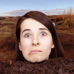 Promotional photo of a white woman with long brown hair. She is frowning theatrically and the photo is edited to make it look like she is buried up to her neck. The background is a brown field and a blue sky.