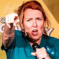 Promotional photo of Pam Ford, a white woman with long red hair. She is speaking energetically into a microphone while pointing at the camera or audience. She is wearing blue scrubs and the background is yellow.