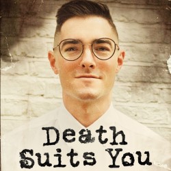 Promotional photo. A white man with short hair and glasses is looking straight ahead at the camera smiling slightly. Black text reads "Death Suits You".