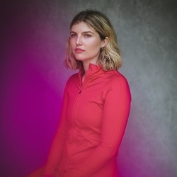 Promotional photo of Jamie-Lee Money, a white woman with blonde hair in a long bob. She is sitting at an angle, looking serious, and wearing a pink exercise top. The bakcground is a greyish lit in gradient purple to green. There is no text.