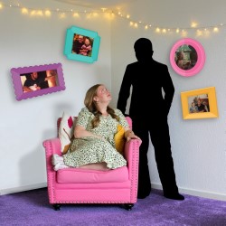 Promotional poster for the play "Strongest Girl in the World". There are two figures, a blonde white woman in a green dress sitting in a childlike way in a pink armchair, and a figure entirely in black, including a black mask covering their face. The walls behind them are covered in colourful picture frames. The text is in yellow childlike writing and reads Strongest Girl in the World. End description.