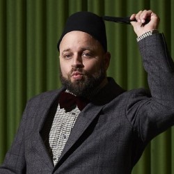 A promotional photo of Victor Esses. He is wearing a grey suit, bow tie and fez, and he is twirling the tassel with one hand raised. He has a quizzical look on his face and he is standing in front of a green curtain.